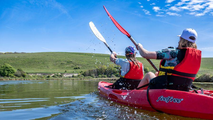 Two people paddling a red kayak on a calm river with rolling green hills in the background.