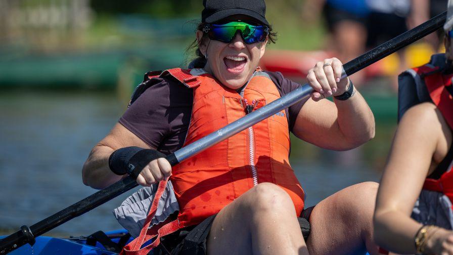 An excited woman in an orange life vest smiling while paddling a kayak.