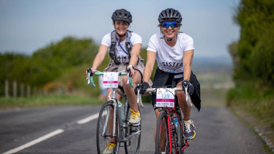 Two women in matching t-shirts smiling and riding road bikes side-by-side up a hill.