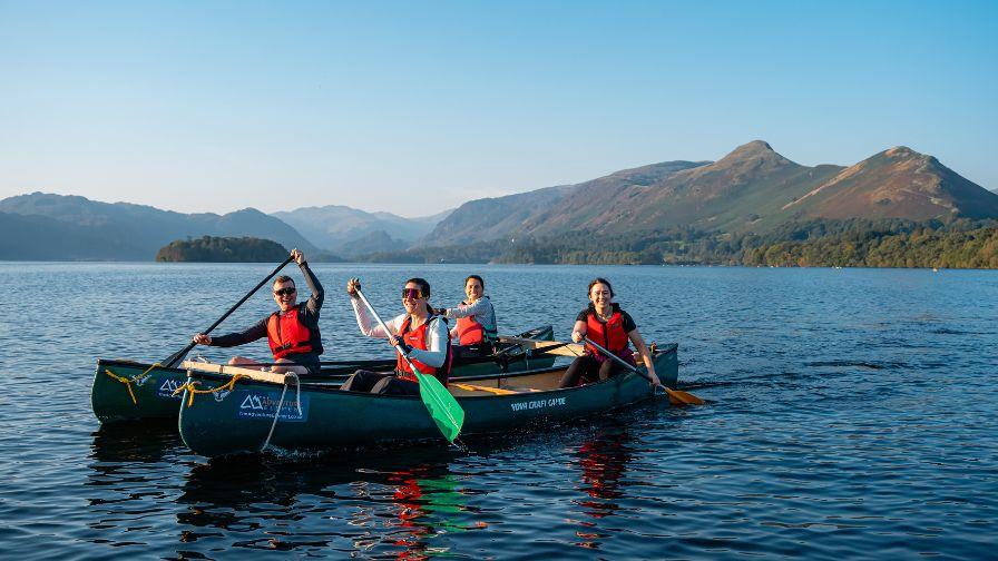 Four people wearing life jackets smiling and paddling in two green canoes side-by-side on a calm lake, with scenic mountains rising in the background under a clear sky.