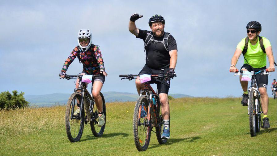 Three mountain bikers smiling and waving while cycling along a scenic grassy ridge.