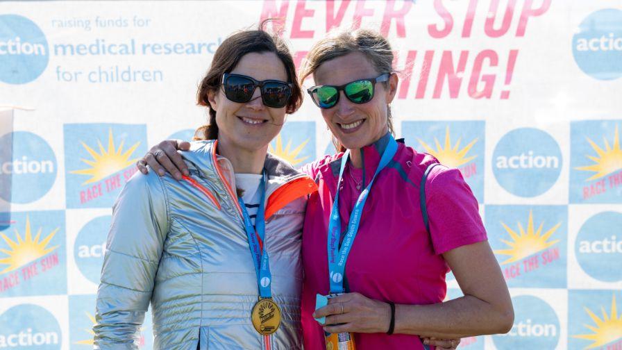 Two women wearing sunglasses and finisher medals smile while posing in front of a charity event banner.