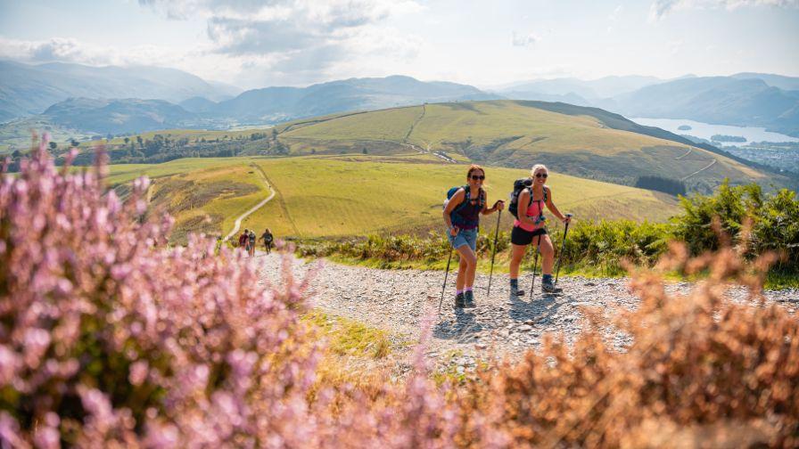 Two women with backpacks and trekking poles hike up a gravel mountain path, smiling as they look ahead, with rolling green hills and a lake visible in the distant landscape.