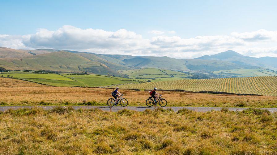 Two cyclists riding along a paved road cutting through a vast, grassy landscape with rolling green hills and mountains in the background under a blue sky with light clouds.