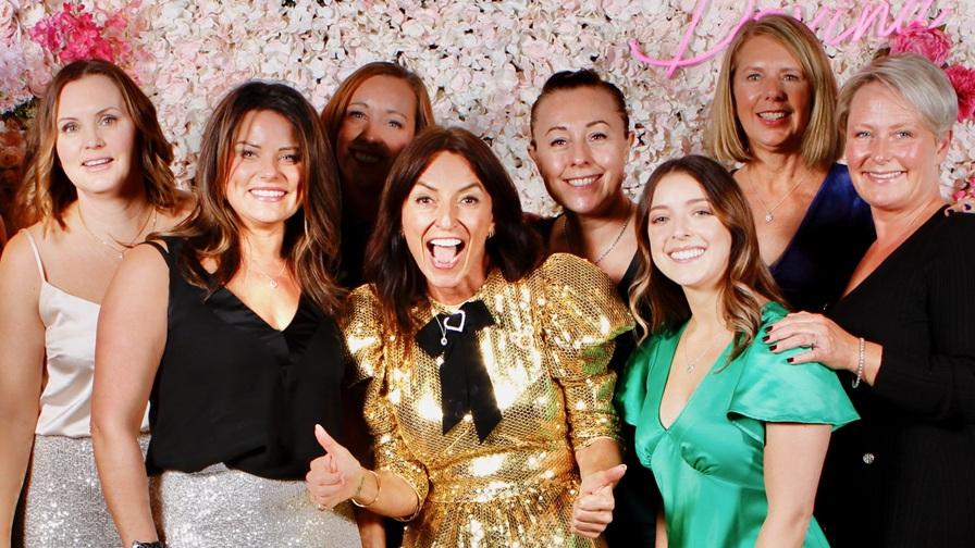 Image shows Davina McCall at the centre of a group of 7 female Dine with Davina event guests. She is wearing a shiny gold dress and giving a thumbs up sign. Everyone is smiling and they are standing in front of a pink floral flower wall backdrop.