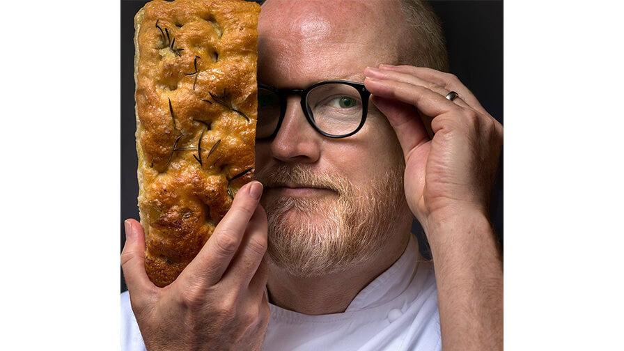 Dan Lepard, a bearded chef adjusting his glasses holds a piece of rosemary focaccia bread up, covering half of his face.