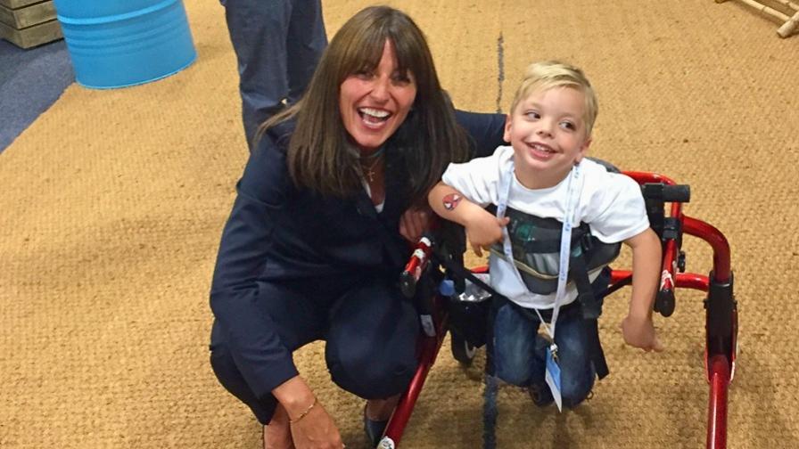 Image shows Davina McCall crouching down next to a little boy who is using a red, wheeled walking frame. He around four years old, has blonde hair and wears a white t-shirt. Both are smiling.