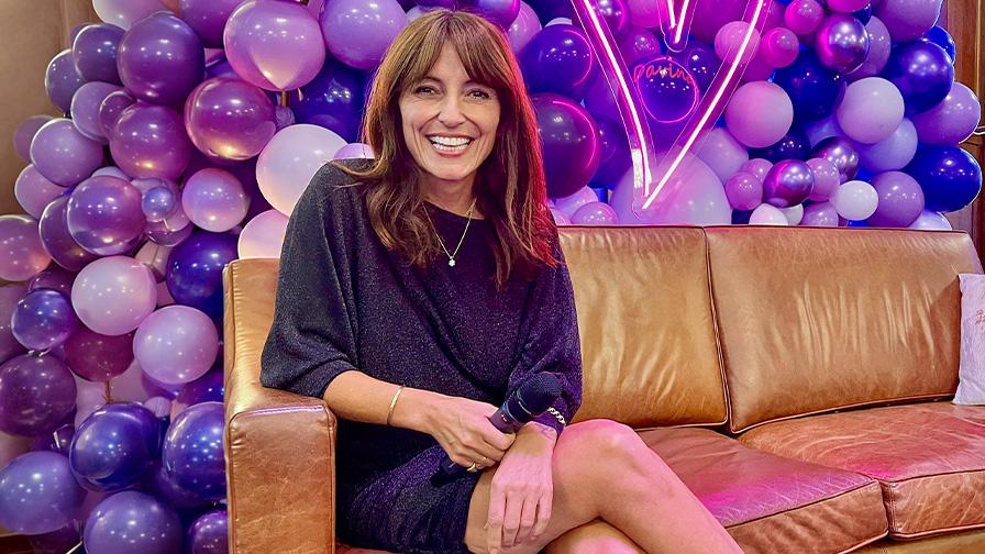 Image shows Davina McCall sitting smiling on a light brown leather sofa. There is a wall of balloons in purples and lilacs behind her.