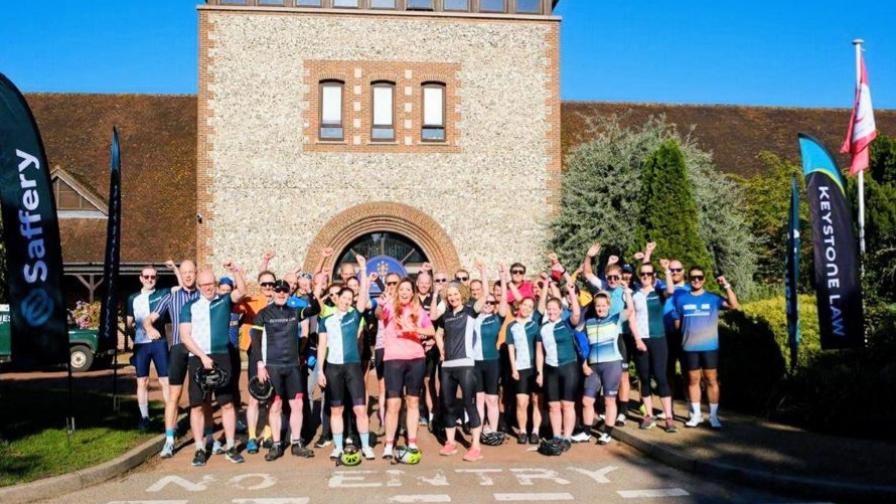 Image shows a large group of people in cycling clothes cheering as they get ready to embark on a charity bike ride.