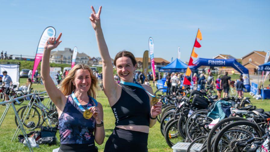 Two happy women holding gold medals at the finish line of race the sun team challenge event.