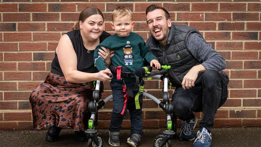 A smiling family poses outdoors against a brick wall with their young son who is standing in a mobile gait trainer.