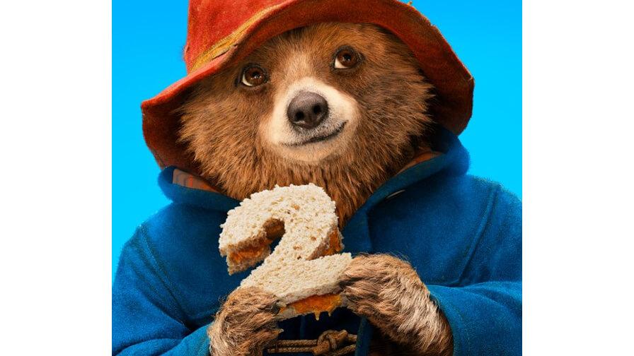Paddington Bear holds a marmalade sandwich shaped like the number 2 against a bright blue background.