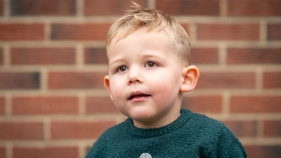 Barnaby, a young boy who has very severe epilepsy. He is earing a dark green jumper and is standing in front of a brick wall.