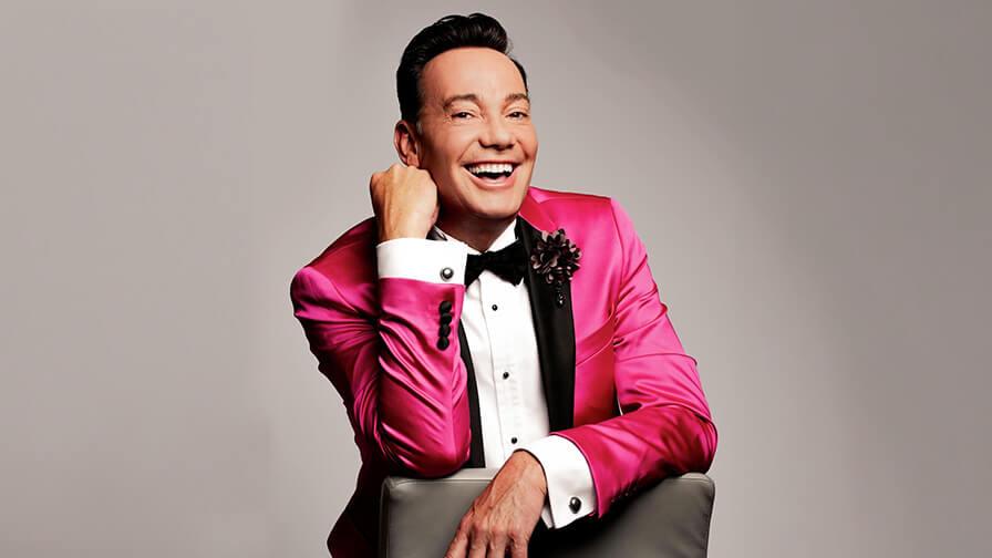 Craig Revel Horwood smiles warmly in a vibrant pink tuxedo jacket and black bow tie, leaning over the back of a chair.