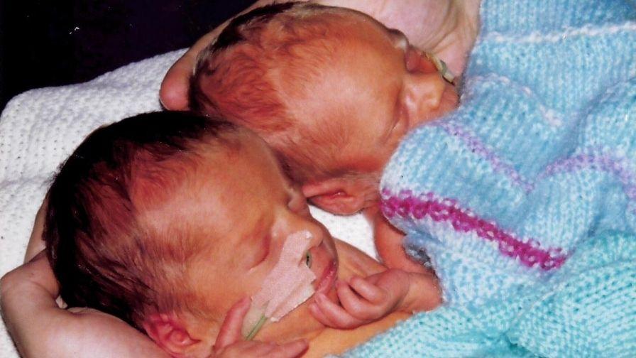 Two tiny premature babies with breathing tubes, laying down cradled in a pair of hands and wrapped in a hand knitted blue blanket