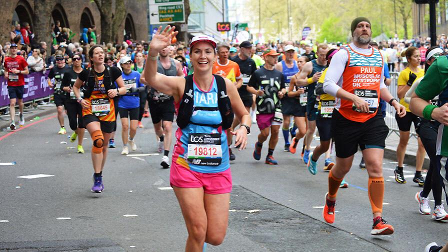A smiling female runner waves at the camera while participating in the London Marathon among a crowd of other runners.
