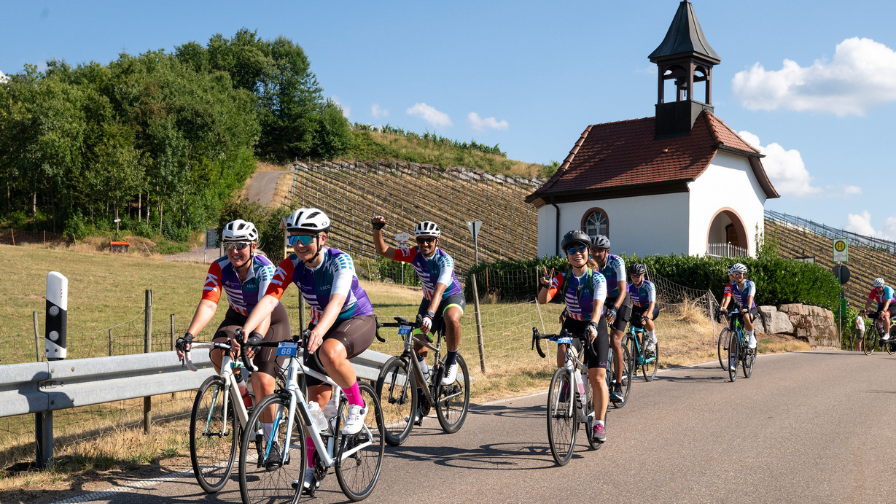 A group of riders cycling along a road in the sunshine; some are waving at the camera and smiling. You can see a small German church behind them. The weather looks hot