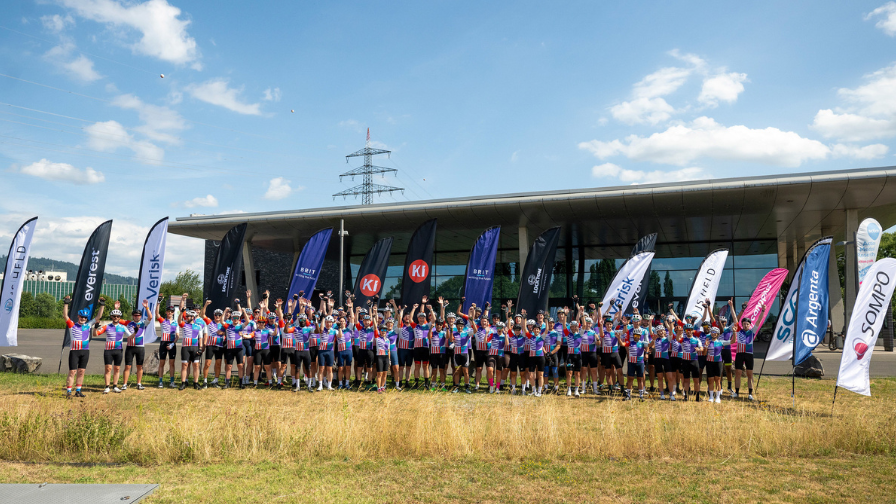 A group of cyclists (around 75) lined up at the start of the ride, waving at the camera for the group shot. The sky behind them is blue