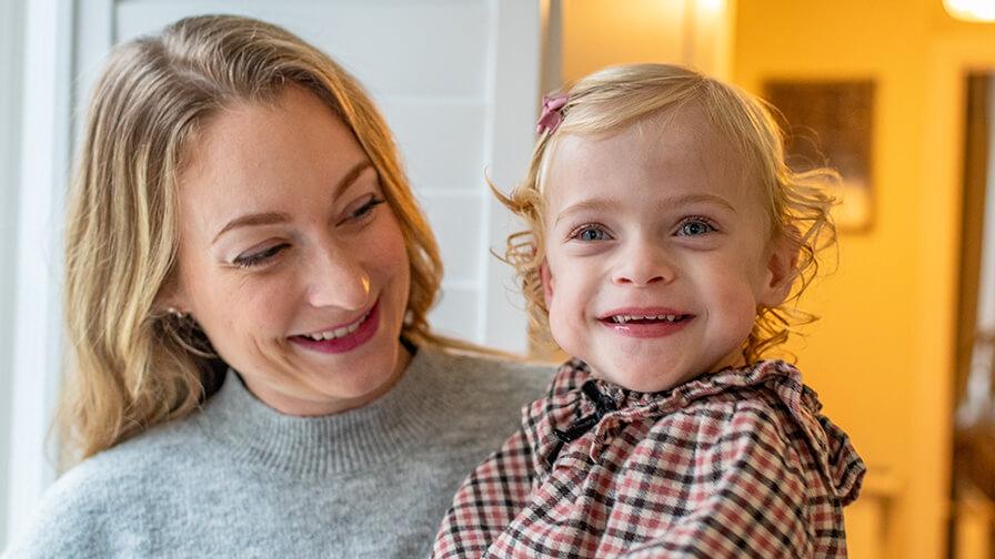 A smiling mother holds her blonde toddler daughter, who wears a plaid dress and smiles at the camera.