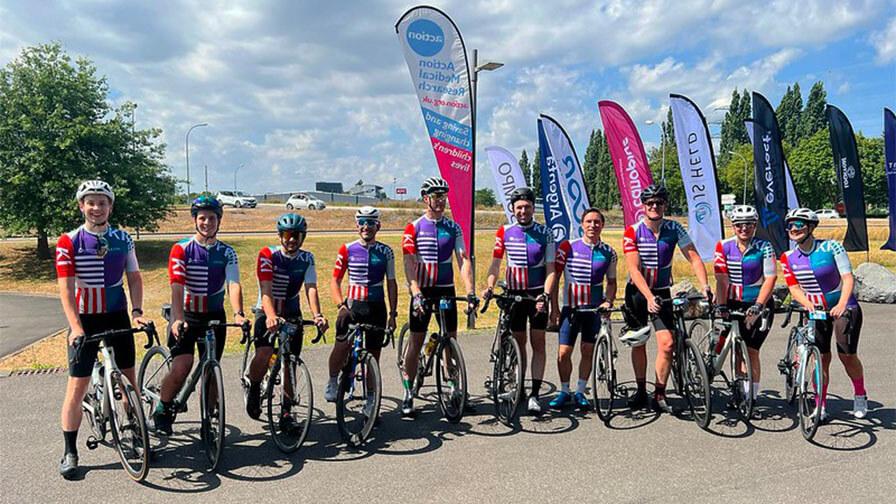 A group of cyclists in matching colorful jerseys pose with their bikes under a sunny sky in front of event flags.