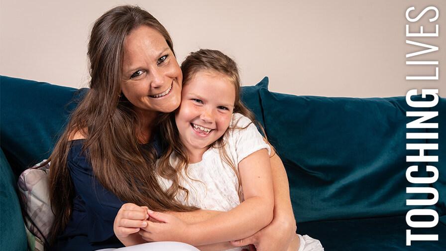 A smiling mother lovingly hugs her young daughter as they sit together on a deep blue velvet sofa.