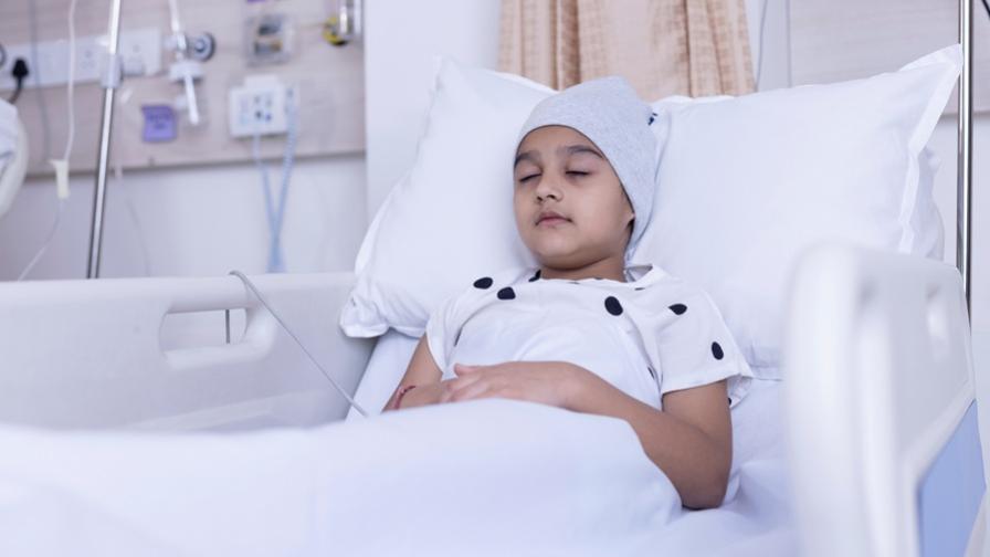 Child in a hospital bed with a bandage around the head, suggesting brain surgery. They are wearing a white gown with black polka dots and resting with their eyes closed.