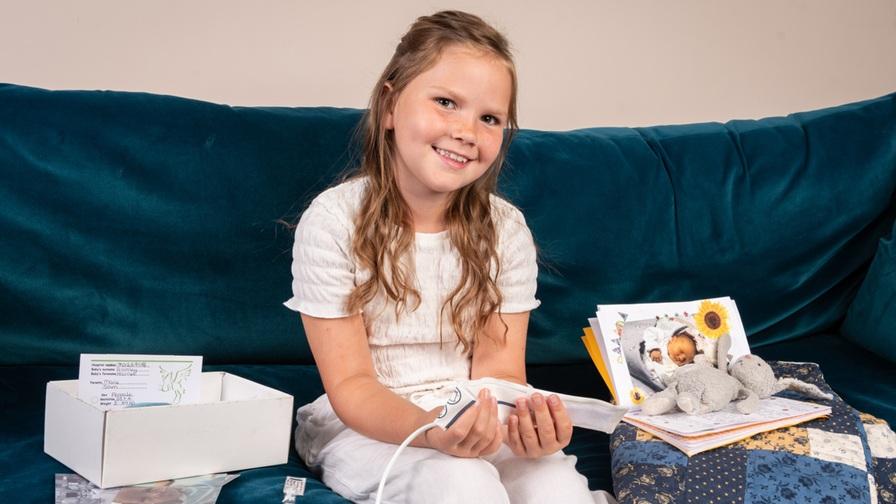 A young girl, Hattie, with long wavy hair sits smiling on a teal velvet sofa. She is holding a medical monitoring device, with an open memory box, printed photographs of a baby, and a small stuffed elephant toy arranged beside her.