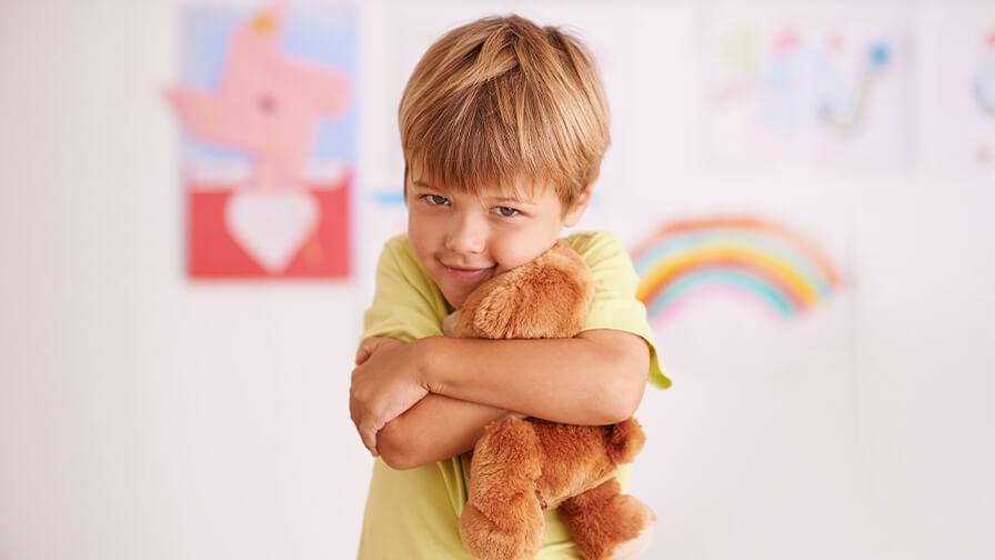 A young boy in a yellow t-shirt smiles as he tightly hugs a brown teddy bear to his chest in a colorful room.