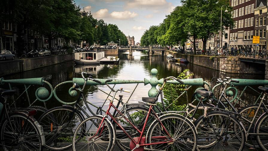 Bicycles parked against a bridge railing along a scenic canal lined with trees and classic architecture in Amsterdam.