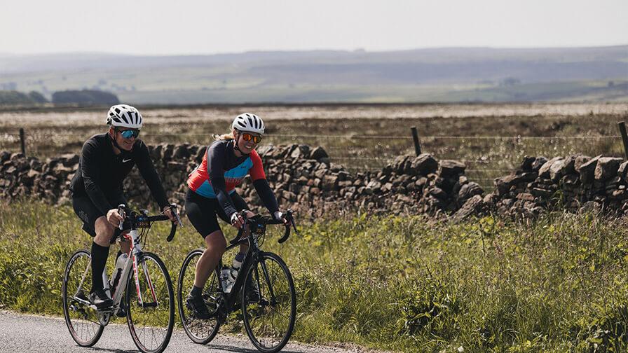 Two cyclists ride road bikes along a rural country lane bordered by a stone wall and open fields.