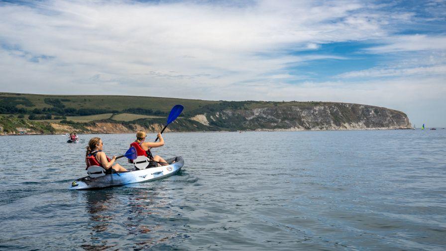 Two people kayaking on calm blue water with white cliffs and green hills in the background.