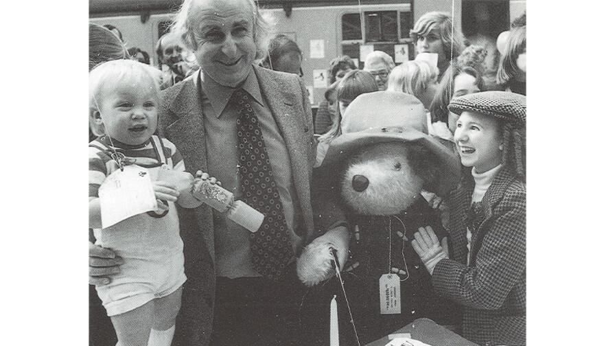 Black and white photo: Muchael Bond, an old man with glasses and a suit, a small kid on his lap, a girl, and a huge plush dog