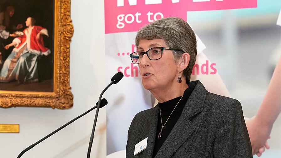 Close-up: Karen Jankel, a woman with glasses and grey hair speaks at a microphone with a painting on the wall