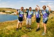 A team of 4 Race the Sun competitors take a moment to pose of picture on rough grassy terain next to a large lake on a sunny day