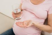 Pregnant woman holding a glass of water in one hand and a supplement tablet in her other outstretched palm.