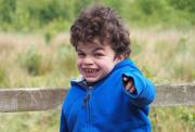  young boy with curly hair wearing a blue jacket, smiling and pointing while standing in front of a wooden fence with greenery in the background.