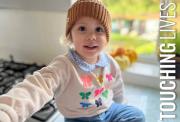 A young child wearing a brown knit hat and butterfly-patterned shirt smiles while sitting in a bright kitchen with pumpkins and plants on the window ledge in the background.