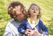 An adult with glasses and messy hair holds a child with a yellow bib in a grassy park, looking at the child with care and concern.