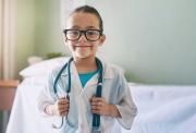 A young girl aged about 7. She wears black-rimmed glasses and is smiling. She is dressed in a white doctor