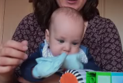 Baby wearing blue sticky mittens, playing with toys.