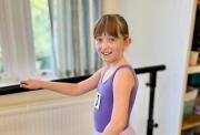 Tora, a 10-year-old girl, standing with her hand on a ballet barre, wearing a purple leotard. 