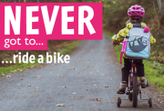 A young child wearing a pink helmet and backpack rides a bike with training wheels on a forest path. Bold text reads, "NEVER got to... ride a bike.