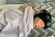 A baby laying asleep in a hospital cot, with a special imaging cap on their head to monitor blood flow in the brain.