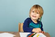 Smiling young child sits at a table holding a green colored pencil, surrounded by blank paper and crayons, ready to draw.