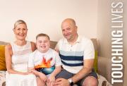 Smiling boy sits on a sofa between a woman in a white dress and a man in a striped polo, posing happily together indoors.