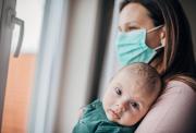 A baby, held by its mother, gazes out at the viewer. The mother is standing by a closed window and wearing a face mask over her nose and mouth.