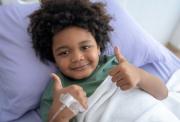 A boy laying in a hospital bed, propped up on a pillow, with an IV drip attached to the back of his hand. He is smiling and giving a two thumbs up sign, symbolising the hope and positive impact of newly funded medical research.