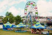  A vibrant summer fair with blue and green tents and a large Ferris wheel under a cloudy blue sky.