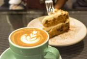Close-up of a latte with leaf art and a slice of cake on a white plate, blurred in the background.