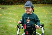 A picutre of Barnaby, a young boy in a green bear sweater and cap smiles while using a supportive walking frame in a grassy garden with fallen leaves